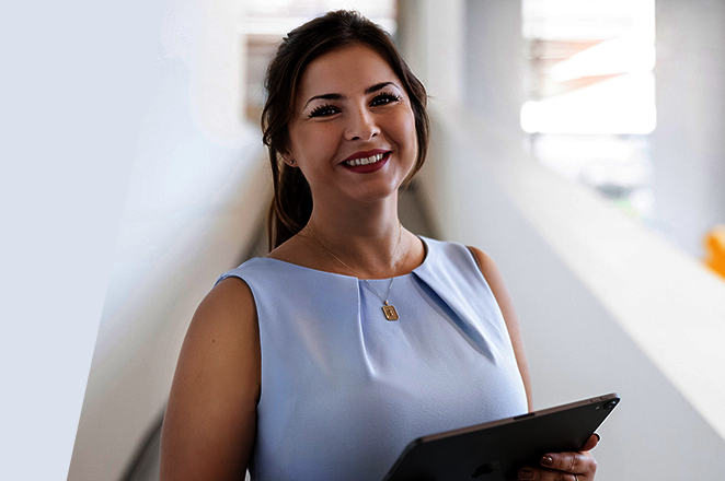 Une femme en robe claire tient une tablette et sourit, tandis qu'elle se tient dans un bureau moderne avec une enseigne d'entreprise visible.