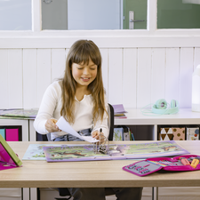 Ein Mädchen sitzt an einem Tisch, lächelt und hält ein Blatt Papier. Auf dem Tisch liegen Bücher und Schulmaterialien.