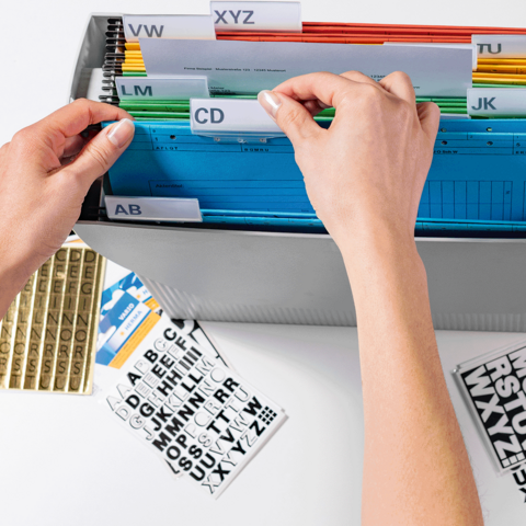 Une main trie des onglets colorés dans un dossier, tandis que des étiquettes avec des lettres sont posées à côté.