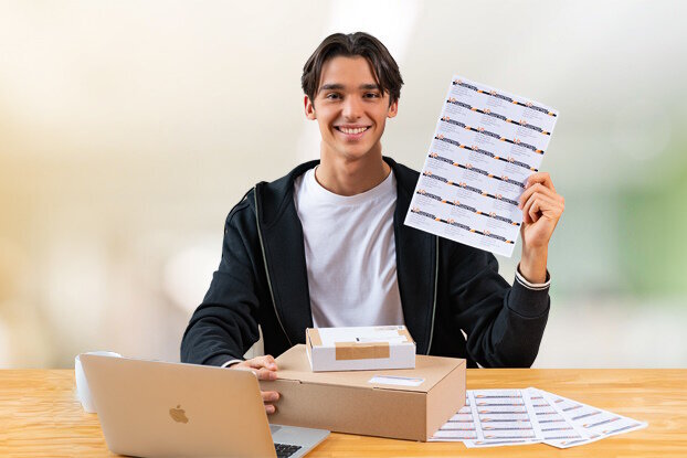 Un jeune homme est assis à une table avec un ordinateur portable et tient une feuille de stickers dans la main.