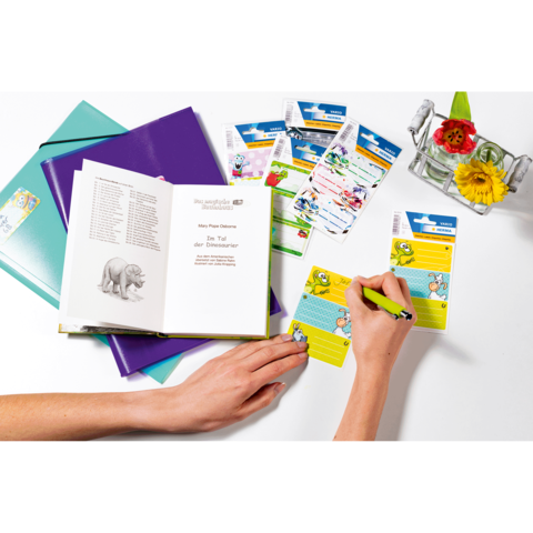Eine Hand schreibt in ein Notizbuch, umgeben von bunten Aufklebern und einem Blumenarrangement. Ein offenes Buch zeigt einen Text über Tiere.