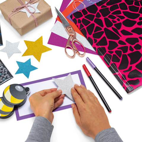 Des mains qui collent une étoile scintillante sur un papier coloré, entourées de fournitures de bricolage comme des ciseaux, des feutres et du papier cadeau.