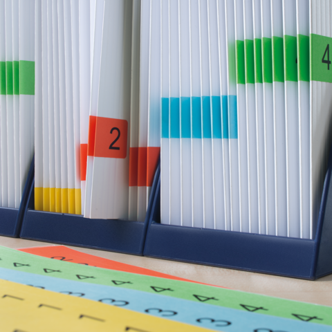 Kartonbox auf einem Tisch mit einem roten Heft und einem grünen "TOP"-Schild. Im Hintergrund sind Bücherregale sichtbar.