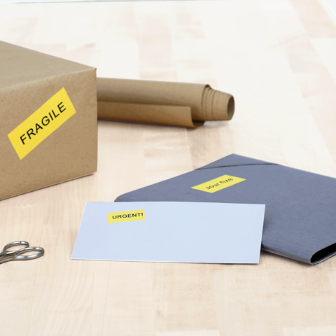 Ein Karton mit dem Aufkleber "FRAGILE", daneben ein graues Kuvert mit der Aufschrift "URGENT!" und eine Schere.