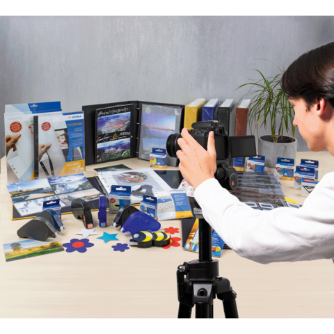 Eine Person fotografiert eine Vielzahl von Büro- und Bastelmaterialien, darunter Ordner, Kleber und kreative Werkzeuge, auf einem Tisch.