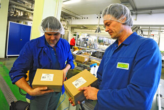 Deux hommes en combinaisons de travail bleues et en filets à cheveux tiennent des cartons dans un environnement de production et vérifient leur contenu.